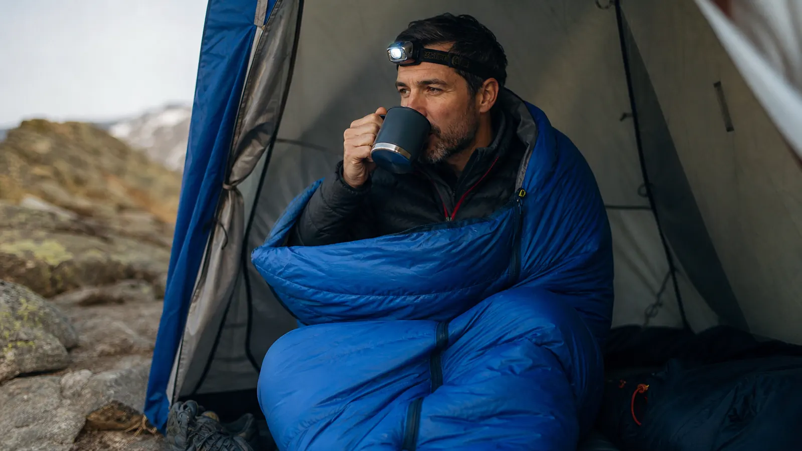 Camper in a mummy sleeping bag inside a tent, wearing a wool hat, reading by headlamp on a cold night