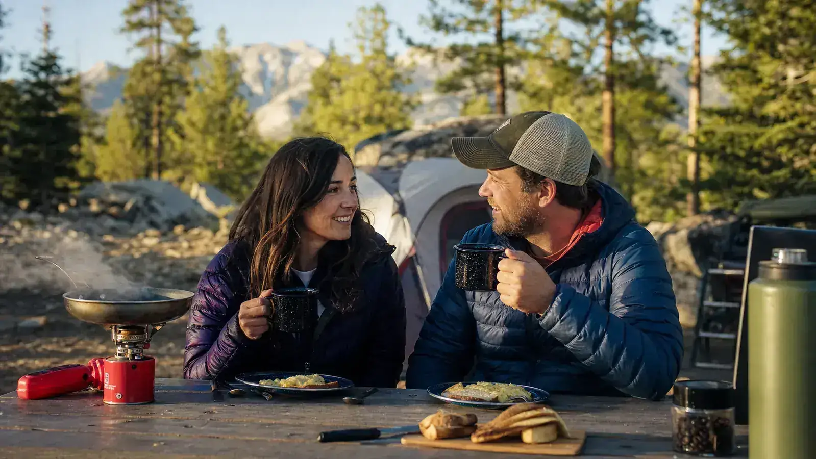 Man and friend eating camp food at a picnic table surrounded by tall pines at dusk